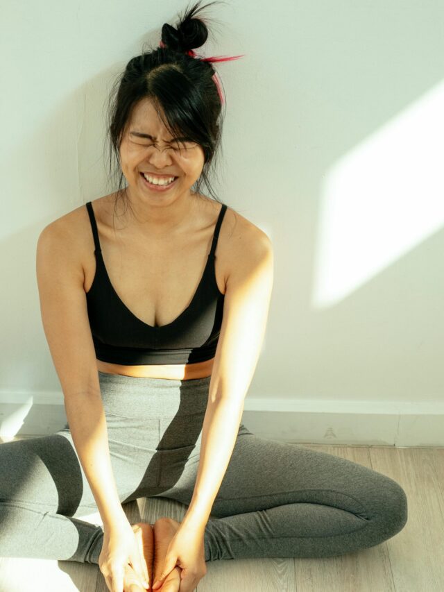 Cheerful young woman practicing butterfly pose on yoga mat in sunny room.